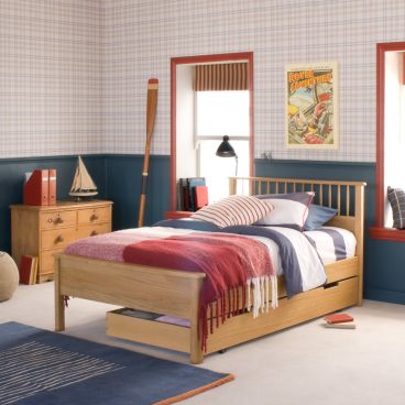 small double oak bed with spindles in teenage boys bedroom. Trundle storage underneath the bed and 2 red framed windows either side 